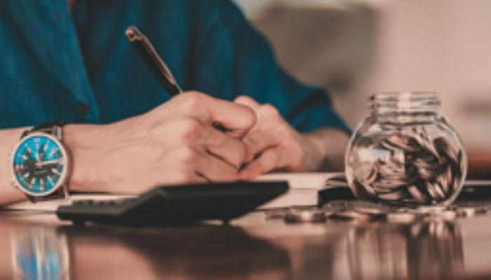 man writing at desk with money jar and calculator