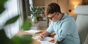 Photo of young student sitting on an office chair and writing at a desk in front of a window in a bright room