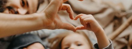 An adult and a child create a heart shape together with their hands, while a baby lies nearby, symbolizing love and family connection.