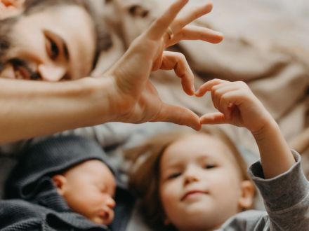 An adult and a child create a heart shape together with their hands, while a baby lies nearby, symbolizing love and family connection.